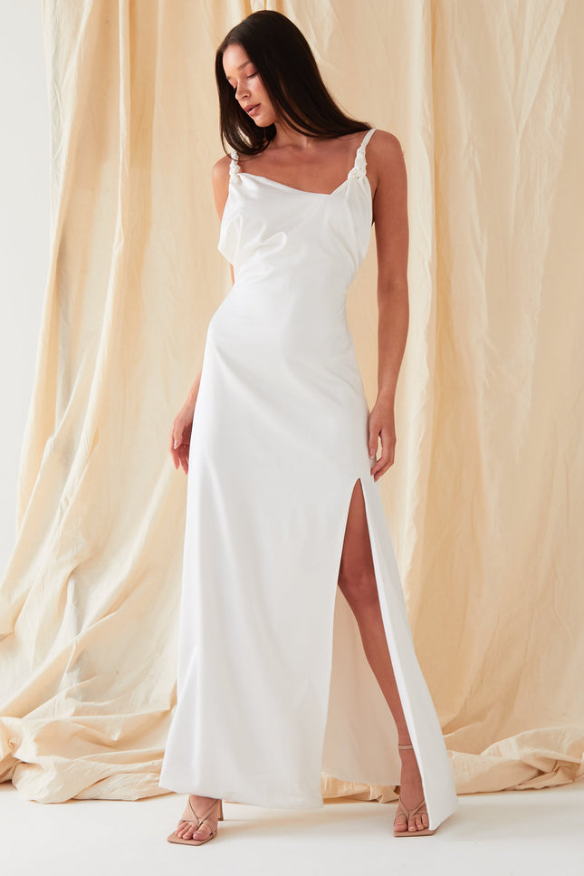 A woman in the Sarvin Ivory Satin Asymmetric Backless Maxi Dress stands against a beige fabric backdrop, wearing strappy heels, her hair down, and gazing downward with effortless elegance.
