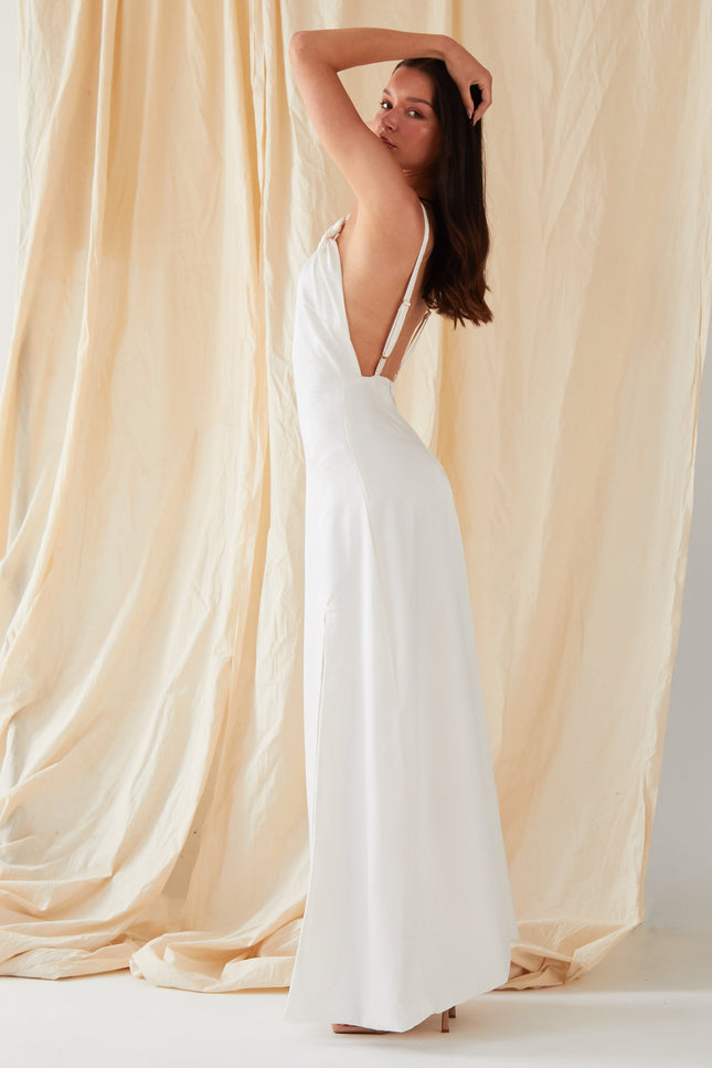 A woman poses in profile, one arm raised, wearing the Sarvin Ivory Satin Asymmetric Backless Maxi Dress with thin straps, set against a beige draped fabric backdrop.