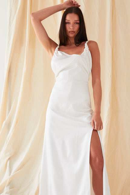 A woman poses before a beige draped backdrop wearing the Sarvin Ivory Satin Asymmetric Backless Maxi Dress, featuring a side slit. She raises one arm above her head and looks directly at the camera.