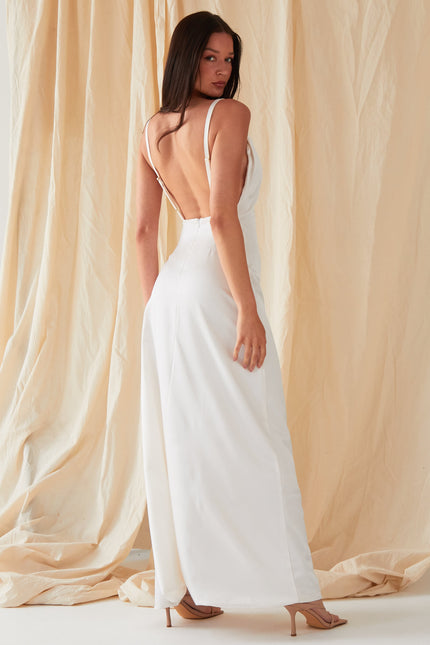 A person with long dark hair wears the Sarvin Ivory Satin Asymmetric Backless Maxi Dress, posing before beige fabric drapes in nude heeled sandals and looking over their shoulder in this elegant evening gown.