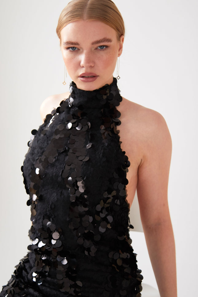 A light-skinned, blonde-haired person with a sleek middle part wears the Sarvin Black Backless Sequin Mini Dress, sitting on a white chair against a plain light background.