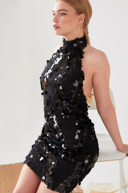 A person with long, light brown hair sits on a white chair, wearing the Sarvin Black Backless Sequin Mini Dress. The dress is covered in large sequins and the background is plain and light-colored.