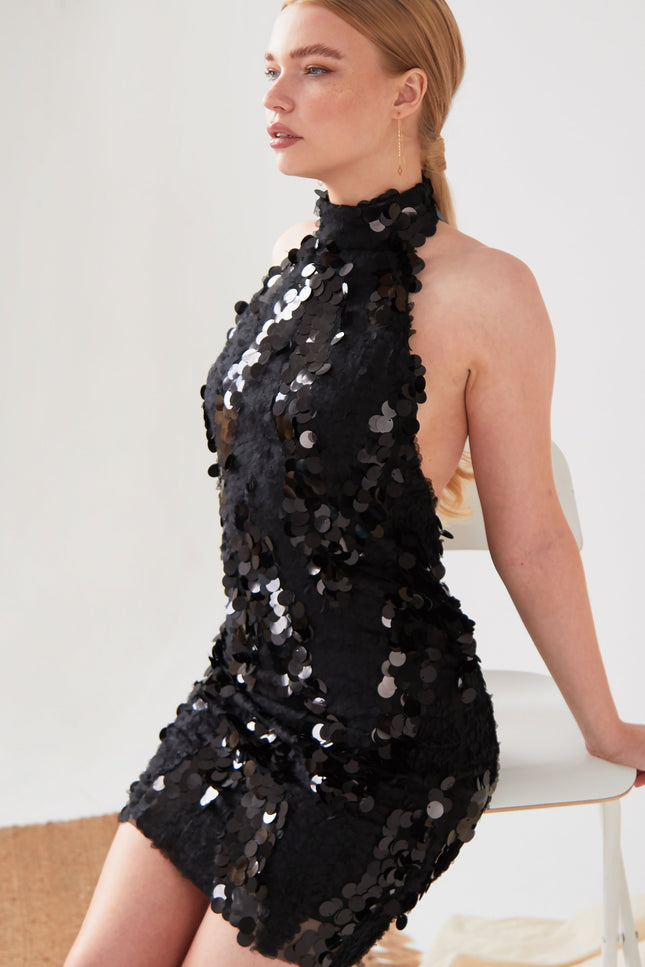 A person with long, light brown hair sits on a white chair, wearing the Sarvin Black Backless Sequin Mini Dress. The dress is covered in large sequins and the background is plain and light-colored.
