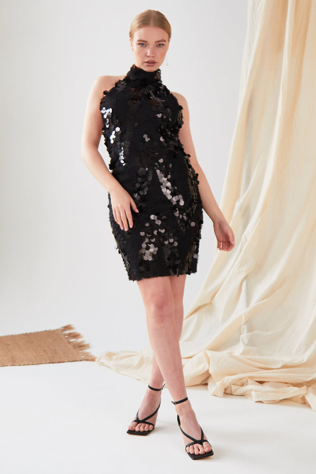 A woman models the Sarvin Black Backless Sequin Mini Dress, standing before cream draped fabric with one leg slightly bent and arms relaxed, paired with black high-heeled sandals.