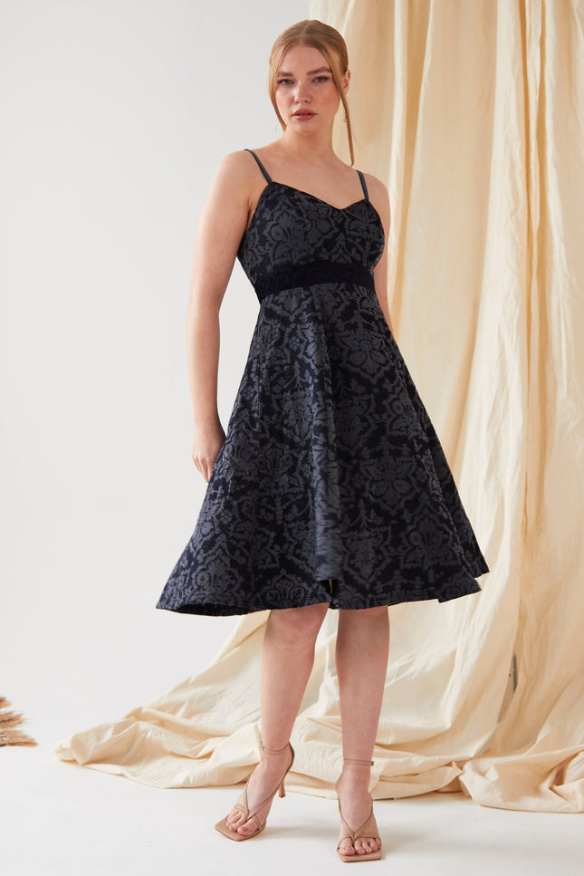 A woman with her hair tied back stands before a beige curtain, wearing the Sarvin Jacquard Skater Dress—navy, sleeveless, knee-length with thin velvet straps and a textured floral pattern—paired with nude high-heeled sandals.