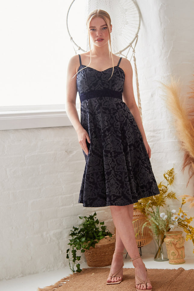 A woman stands indoors in front of a white brick wall, surrounded by plants and dried grasses, wearing the Sarvin Jacquard Skater Dress with velvet straps and strappy heels.