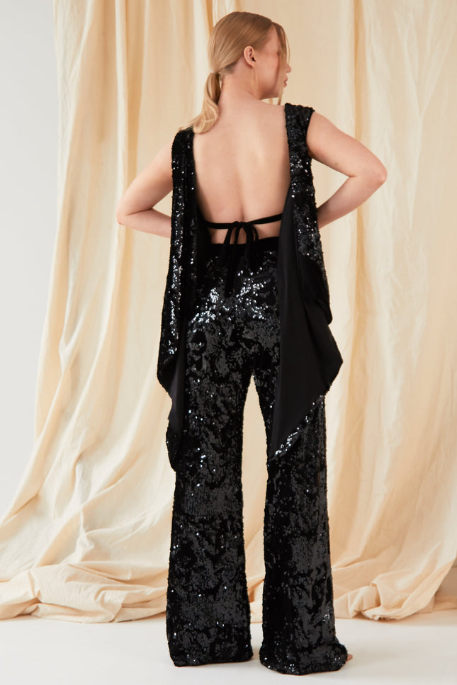 A person faces away from the camera, wearing Sarvin’s handmade Sequin Velvet Flared Trousers and a matching backless sequin top, posed before light beige draped fabric.