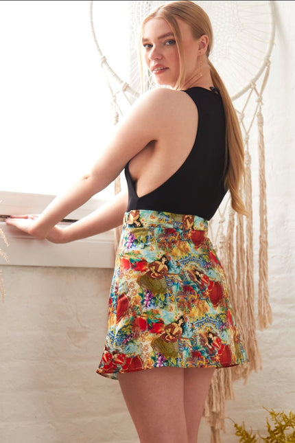 A woman with long blonde hair stands by a window in a sleeveless black top and the Sarvin Persian Printed Mini Skirt, looking over her shoulder at the camera. The background features dried plants and a macramé wall hanging.