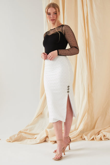 A woman stands before a beige draped backdrop, wearing the Sarvin Long-Sleeve Mesh Bodysuit, a white textured midi skirt with side buttons and slit, and nude high-heeled sandals, looking toward the camera.