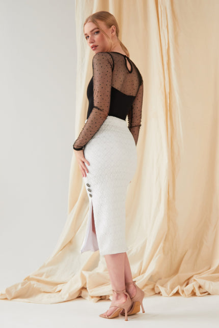 A woman poses before a beige fabric backdrop wearing the Sarvin Long-Sleeve Mesh Bodysuit with glitter polka dots, a white midi skirt with side slit and buttons, and nude heeled sandals, glancing over her shoulder.