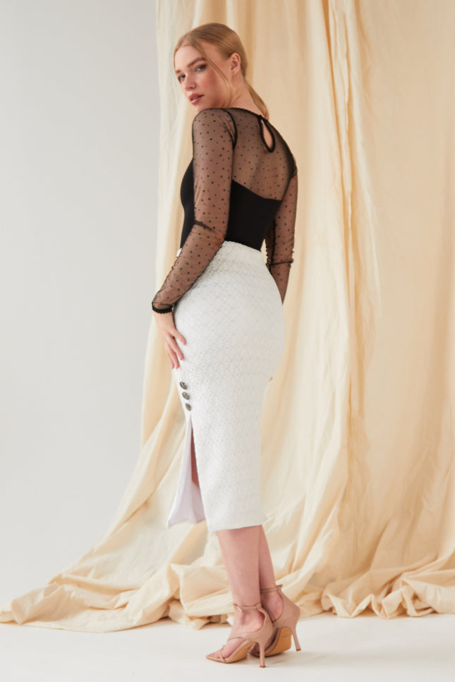 A woman poses before a beige fabric backdrop wearing the Sarvin Long-Sleeve Mesh Bodysuit with glitter polka dots, a white midi skirt with side slit and buttons, and nude heeled sandals, glancing over her shoulder.