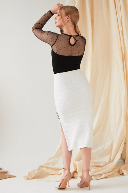 A woman faces away, wearing a black sheer polka dot top and the Sarvin White Bodycon Lace Skirt—handmade in the UK with a high waist, textured fabric, and side slit—paired with nude heels against a cream draped background.