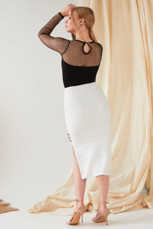 A woman faces away, wearing a black sheer polka dot top and the Sarvin White Bodycon Lace Skirt—handmade in the UK with a high waist, textured fabric, and side slit—paired with nude heels against a cream draped background.