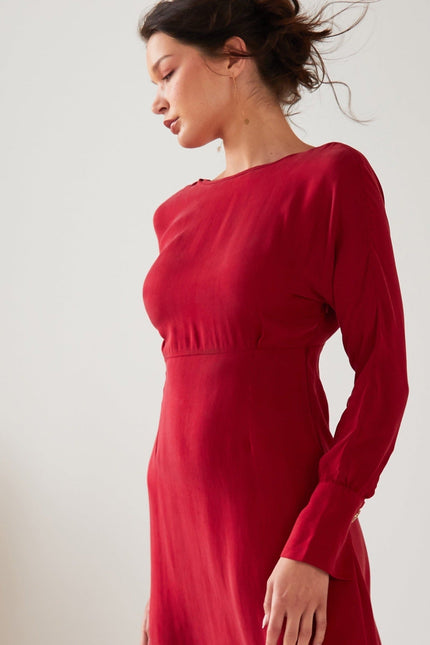 A woman with dark hair tied up stands in profile wearing the Sarvin Long Sleeve Red Midi Dress. The backdrop is plain and light, with part of a plant on the left.