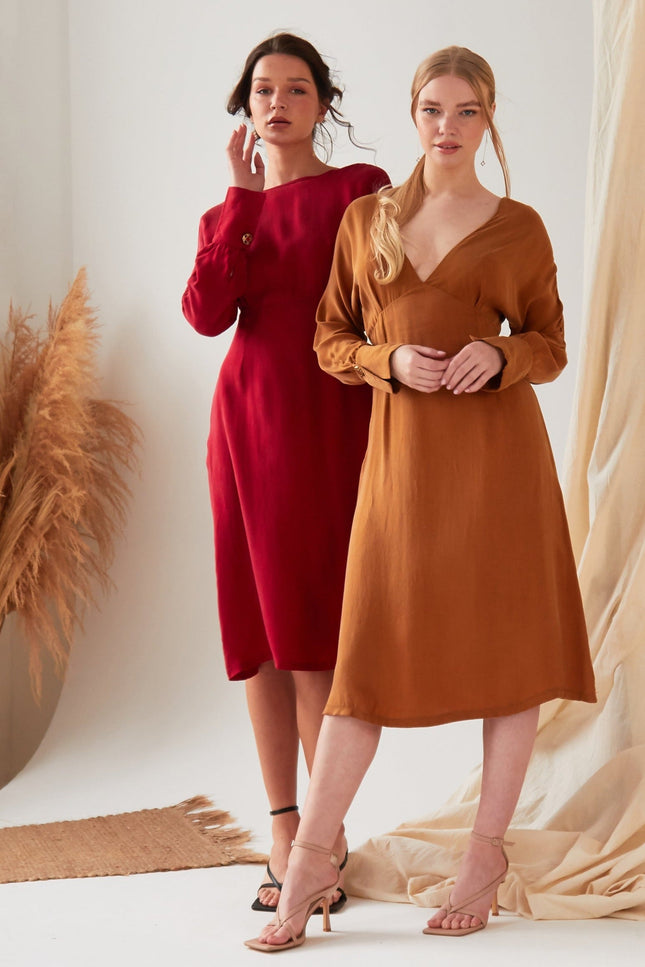 Two women pose confidently in a minimal studio with pampas grass and draped fabric. One wears the Sarvin Mustard Yellow Midi Dress, while the other sports a brown long-sleeve v-neck style; both pair their looks with heels.