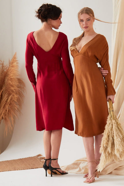 Two women indoors stand amid neutral drapes. One, facing away, wears the Sarvin Long Sleeve Red Midi Dress with black heels; the other faces forward in a brown eco-friendly Cupro dress and holds pampas grass.