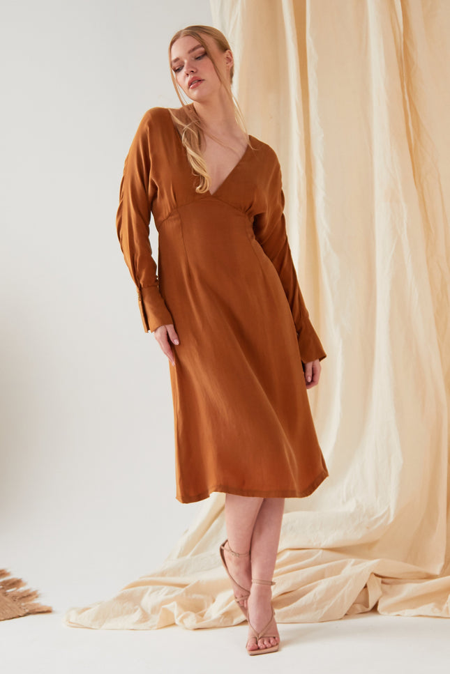 A person wearing the Sarvin Mustard Yellow Midi Dress with long sleeves and a V-neckline, styled with strappy sandals, stands before draped beige fabric.