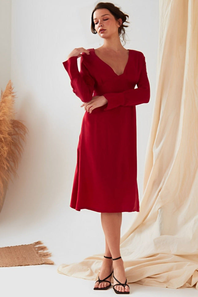 A woman wearing the Sarvin Long Sleeve Red Midi Dress stands indoors near beige curtains and a vase of pampas grass.