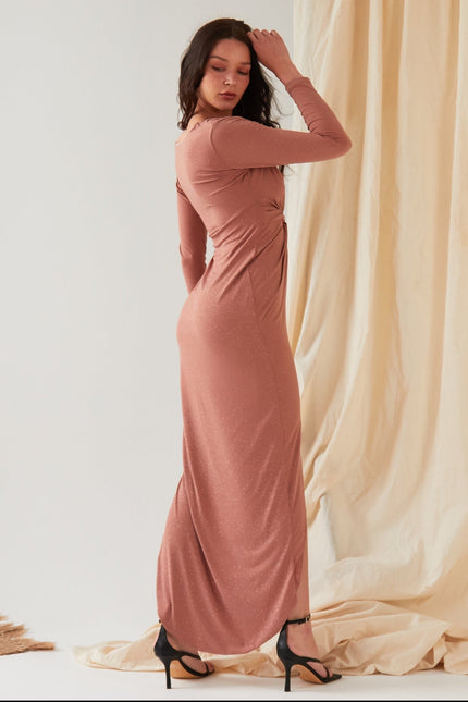 A woman stands sideways against draped fabric, wearing the Sarvin Rose Gold Twist Knot Front Dress with long sleeves and a fitted silhouette, paired with black high-heeled sandals and one hand touching her head.
