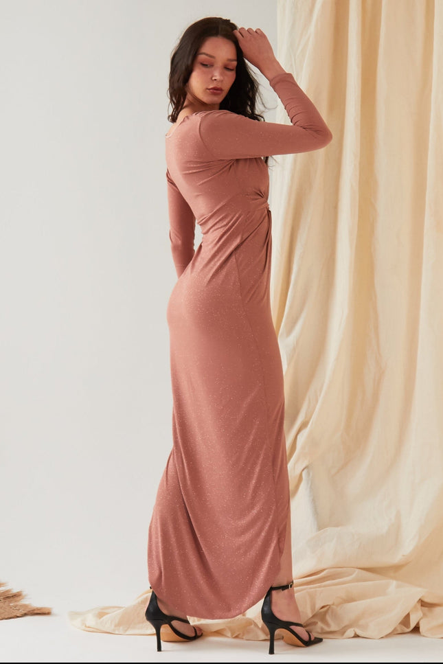 A woman stands sideways against draped fabric, wearing the Sarvin Rose Gold Twist Knot Front Dress with long sleeves and a fitted silhouette, paired with black high-heeled sandals and one hand touching her head.