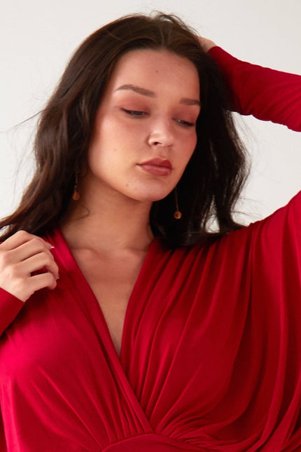 A woman with long dark hair wears Sarvin’s Circle Shape 18K Gold Drop Earrings and a red, long-sleeved draped dress. Posing against a plain light background, she gazes down with one hand near her head and the other at her shoulder.