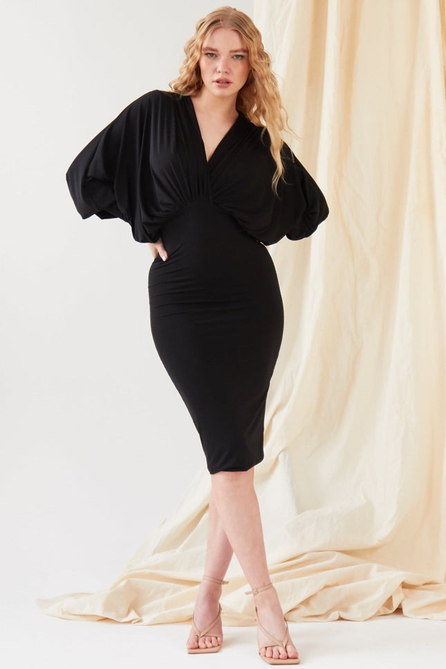 A woman poses before a beige draped backdrop in the Sarvin Black Batwing Sleeve Midi Dress, featuring silk-touch jersey. She pairs the look with nude-heeled sandals and lets her long wavy blonde hair frame her neutral expression.