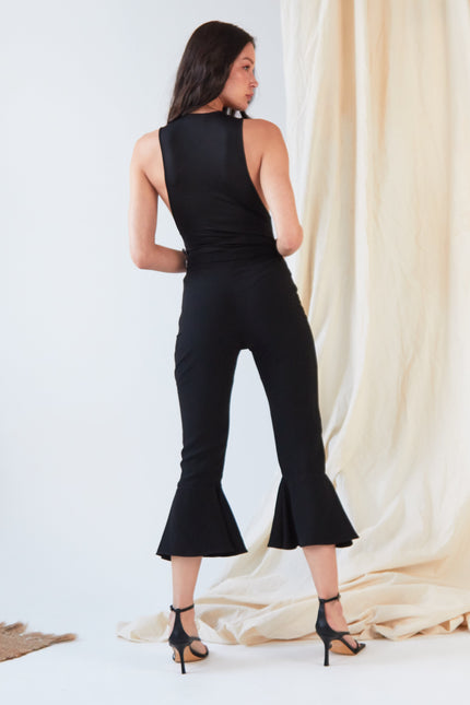 A woman stands facing away, wearing the Sarvin Dropped Armhole Bodysuit with flared hems and black high-heeled sandals. A beige draped fabric hangs in the background.