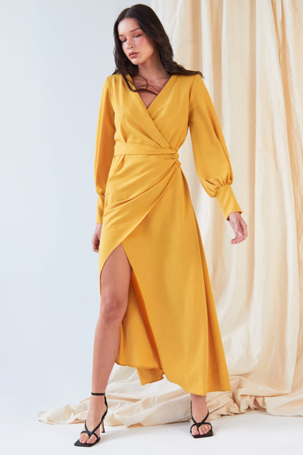 A woman poses before cream drapes in the Sarvin Mustard Wrap Maxi Dress, featuring vintage-inspired satin, balloon sleeves, a deep V-neck, and an asymmetrical hem, styled with black strappy high-heeled sandals.