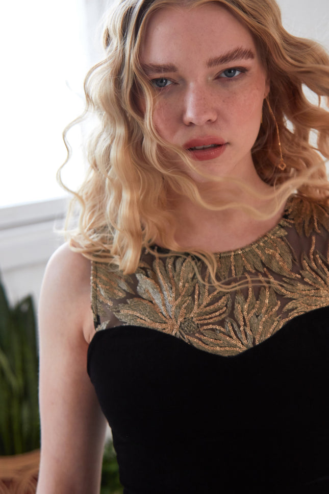 A woman with wavy blonde hair wears a black dress with a sheer, gold-embroidered neckline and Sarvin Clover Shape 18K Gold Threader Earrings. She stands indoors with plants in the background, looking at the camera.