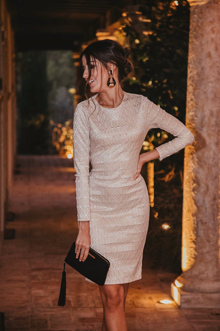 A woman poses indoors at night in the Sarvin Ivory Lace Backless Dress, holding a black clutch with one hand on her hip. Warm lighting and columns accent the background.