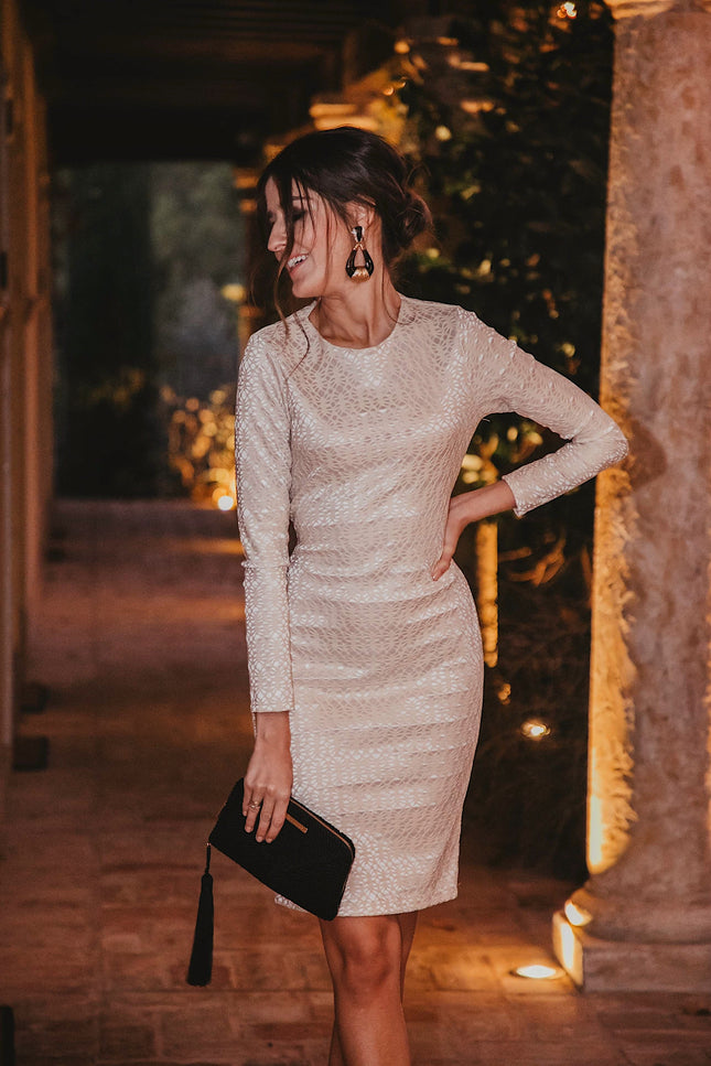A woman poses indoors at night in the Sarvin Ivory Lace Backless Dress, holding a black clutch with one hand on her hip. Warm lighting and columns accent the background.