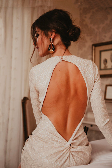 A woman with dark hair in an updo sits on a bed in a softly lit room, wearing the Sarvin Ivory Lace Backless Dress, its delicate open-back detail elegantly highlighted as she faces away from the camera.