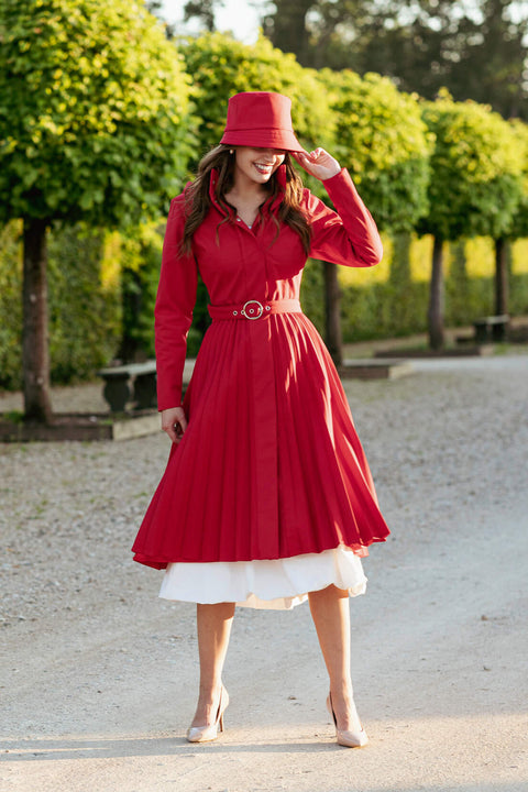 Woman in a red coat and hat standing outdoors with greenery in the background
