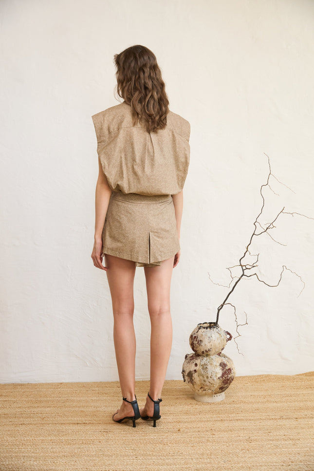 Woman in beige outfit standing with back to camera, vase on floor.