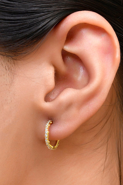 A close-up image of a woman's ear wearing a gold-colored hoop earring studded with cubic zirconia stones.