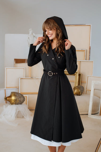 A woman smiling and posing in a double-breasted black trench coat with a hood and a black ruffle belt with silver-tone hardware.