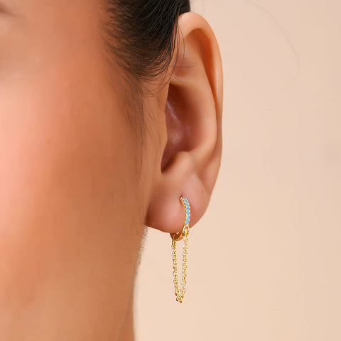 A person wearing a turquoise stone linked huggie earring with a gold plated chain detail.