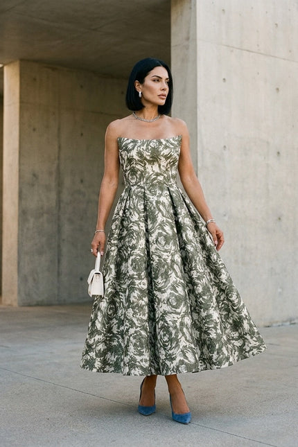 A woman stands outdoors against a concrete wall, wearing the Sarvin Tala Corset Midi Dress, styled with straight black hair, jewelry, blue high heels, and holding a small white purse.
