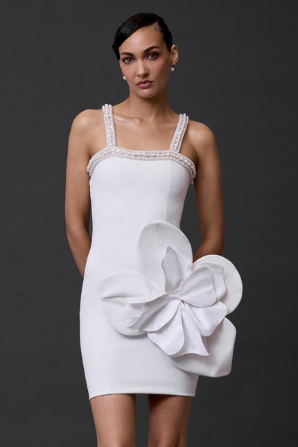 A woman standing and wearing a white dress with a floral pattern, consisting of a sophisticated bow detail on the waist.