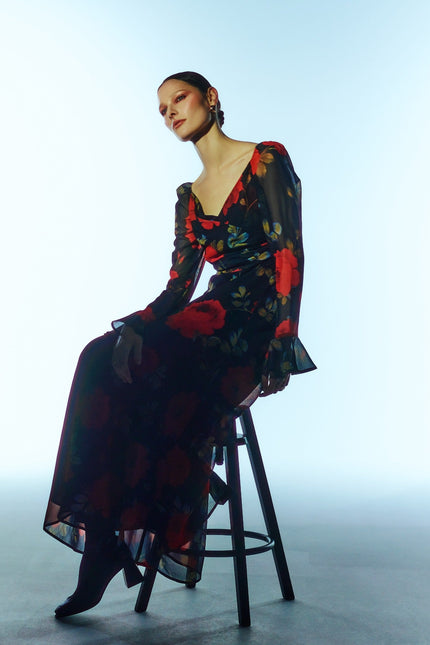 Seated three-quarter model view of the SOUR FIGS Floral Long-Sleeve Maxi Dress in Midnight Rose Black, with red roses, a ruched V-neck bodice and sheer sleeves with flared cuffs.