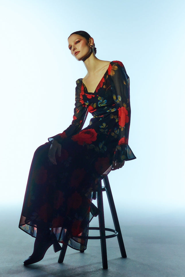 Seated three-quarter model view of the SOUR FIGS Floral Long-Sleeve Maxi Dress in Midnight Rose Black, with red roses, a ruched V-neck bodice and sheer sleeves with flared cuffs.