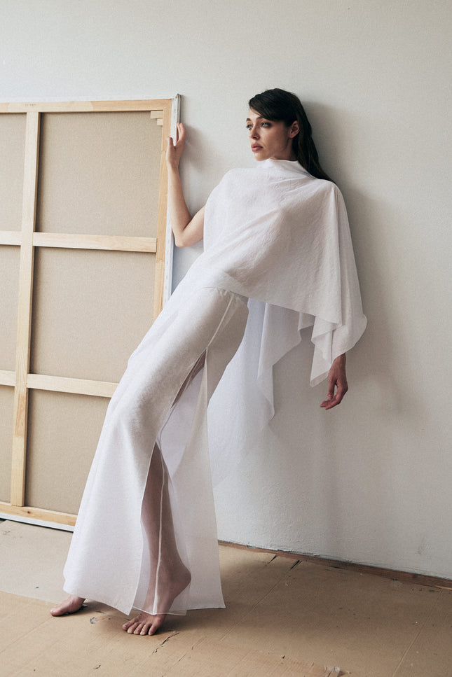 Woman in a white outfit standing next to a large wooden frame against a plain wall.