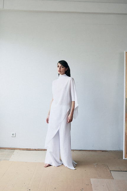 Woman in a white outfit standing in a minimalistic room with a plain wall and wooden floor.