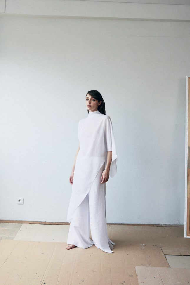 Woman in a white outfit standing in a minimalistic room with a plain wall and wooden floor.