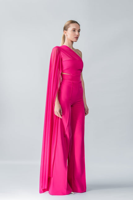 Model wearing a bright pink one-shoulder top and trouser  set with a draped effect on a gray background