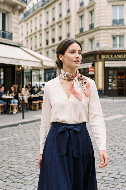 Woman wearing a delicate watercolor floral silk scarf tied around her neck, styled with an elegant cream blouse and navy skirt in a Parisian street setting.