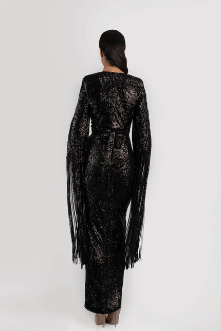 Girl wearing a black sequin job dress with fringe sleeves, backing the camera and  posed against a light grey background.