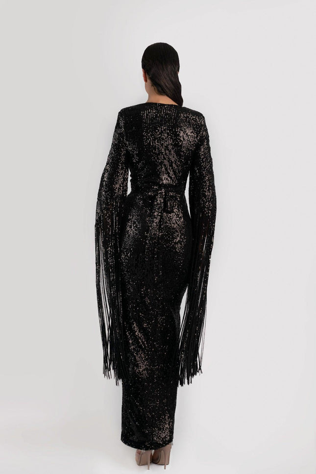 Girl wearing a black sequin job dress with fringe sleeves, backing the camera and  posed against a light grey background.