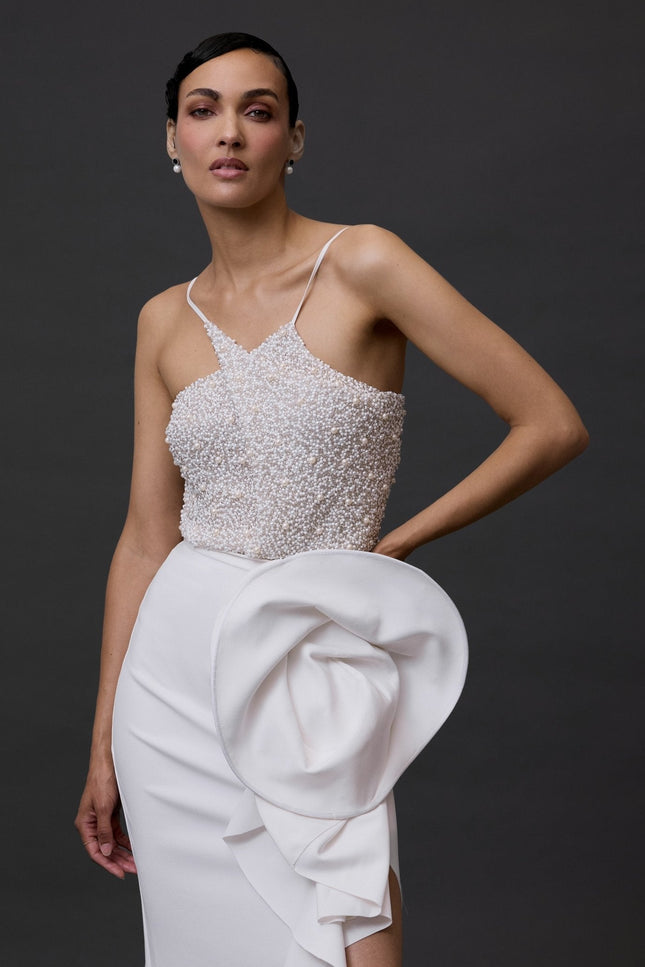 A woman wearing a white beaded bodysuit and a structured skirt, standing against a black background.