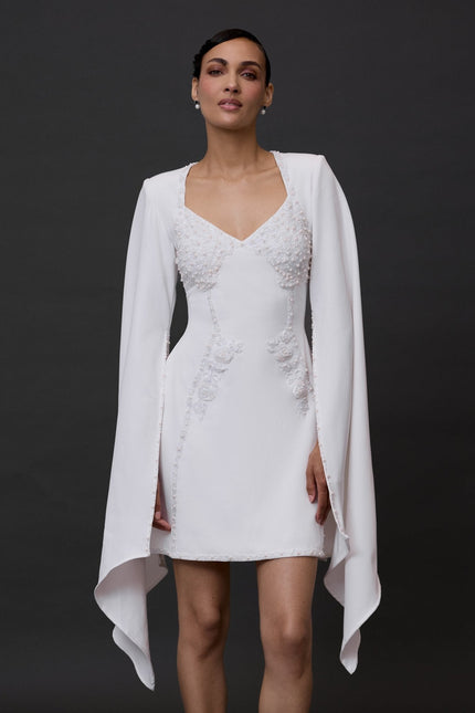 A woman modeling a white mini dress with long sleeves and a V-neckline, featuring a flowing skirt.